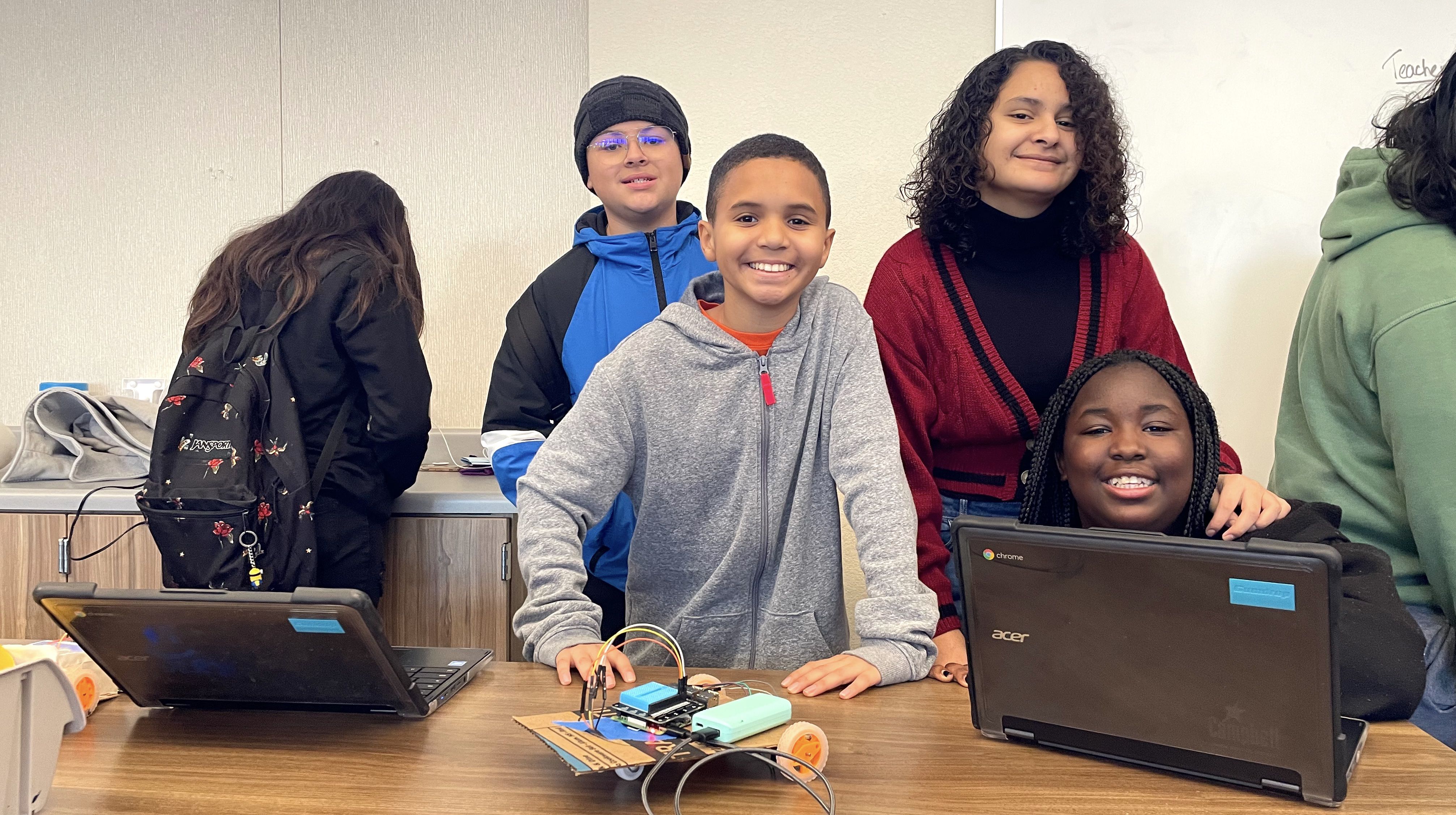 four middle school students smile beside their laptop and hand-made and coded robot vehicle