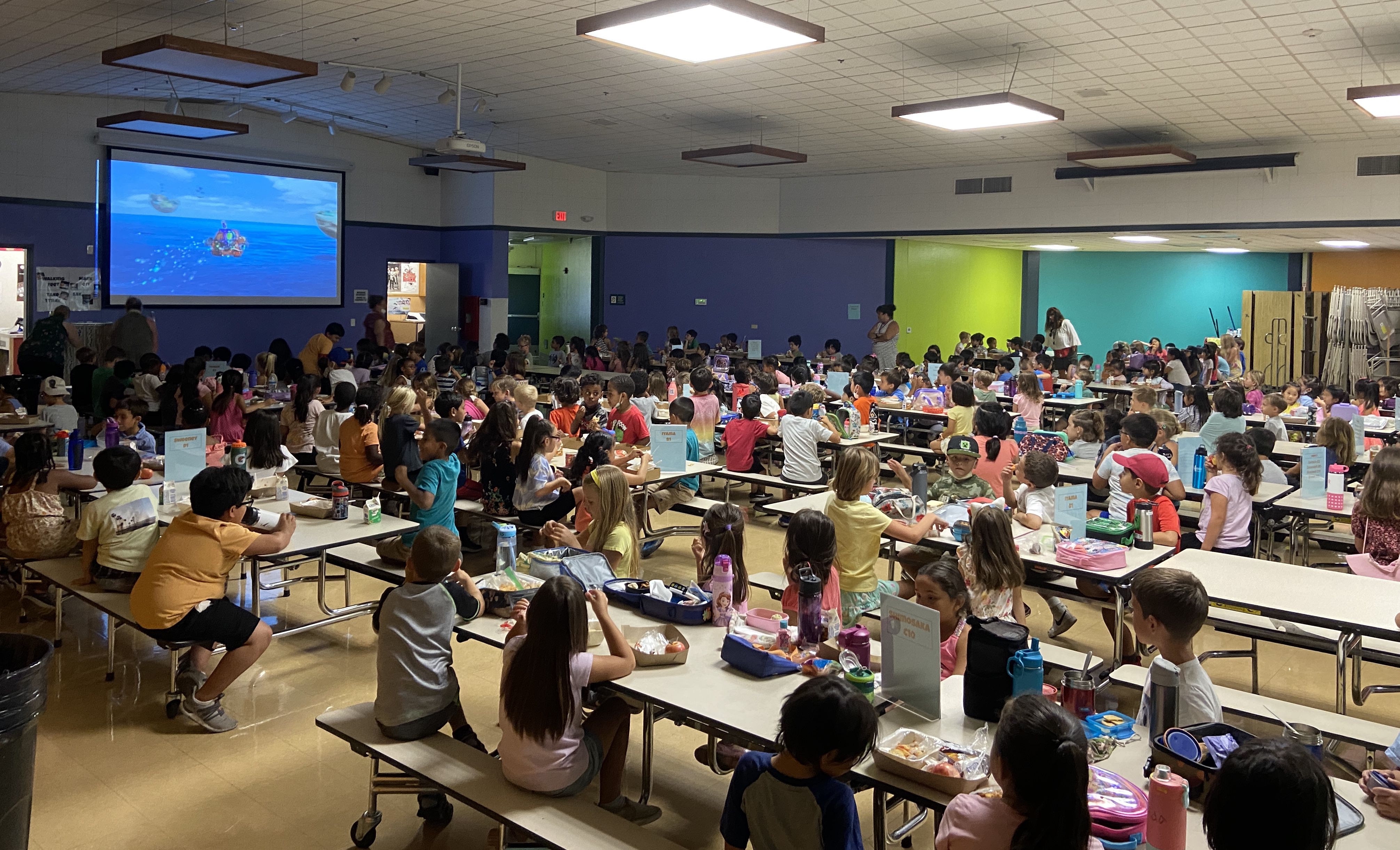 cafeteria filled with young students watching a video on large screen