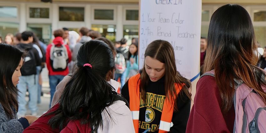 middle school girl wearing an orange safety vest stands at a participation sign up station as several students stand waiting to sign up