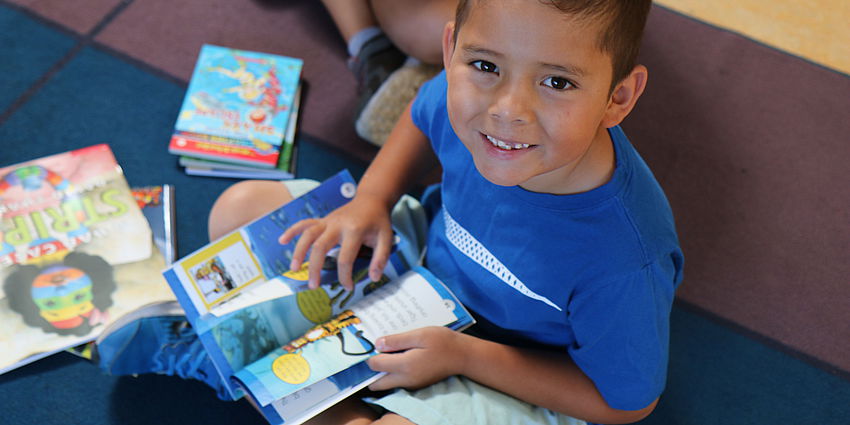 young boy smiling up at camera as he sits on the floor with a book