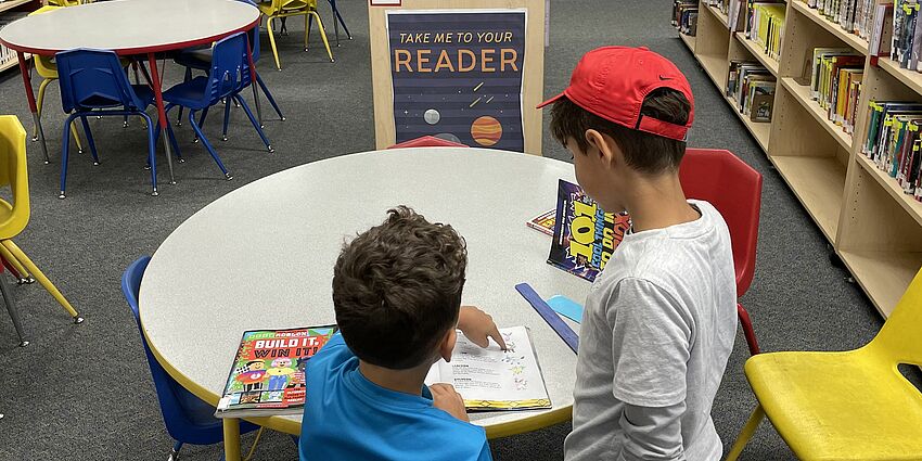 two boys in library in front of a sign that says Take Me To Your Reader