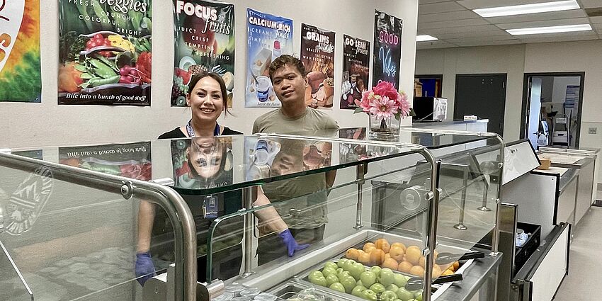 man and woman stand behind cases of fresh food in school lunch line