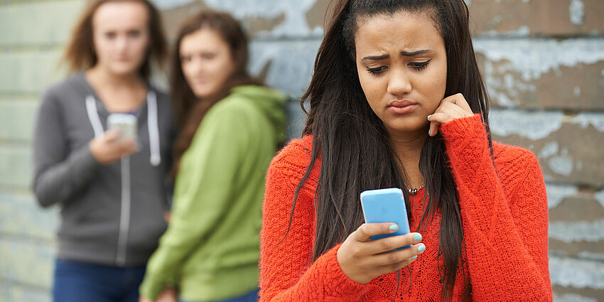 unhappy adolescent girl looking at her phone as two others behind her look and laugh at her