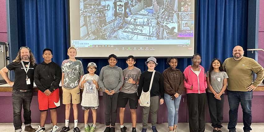 9 Zero Robotics students and 2 adult advisors stand before a screen telecast of the ISS as an astronaut tests the team's code