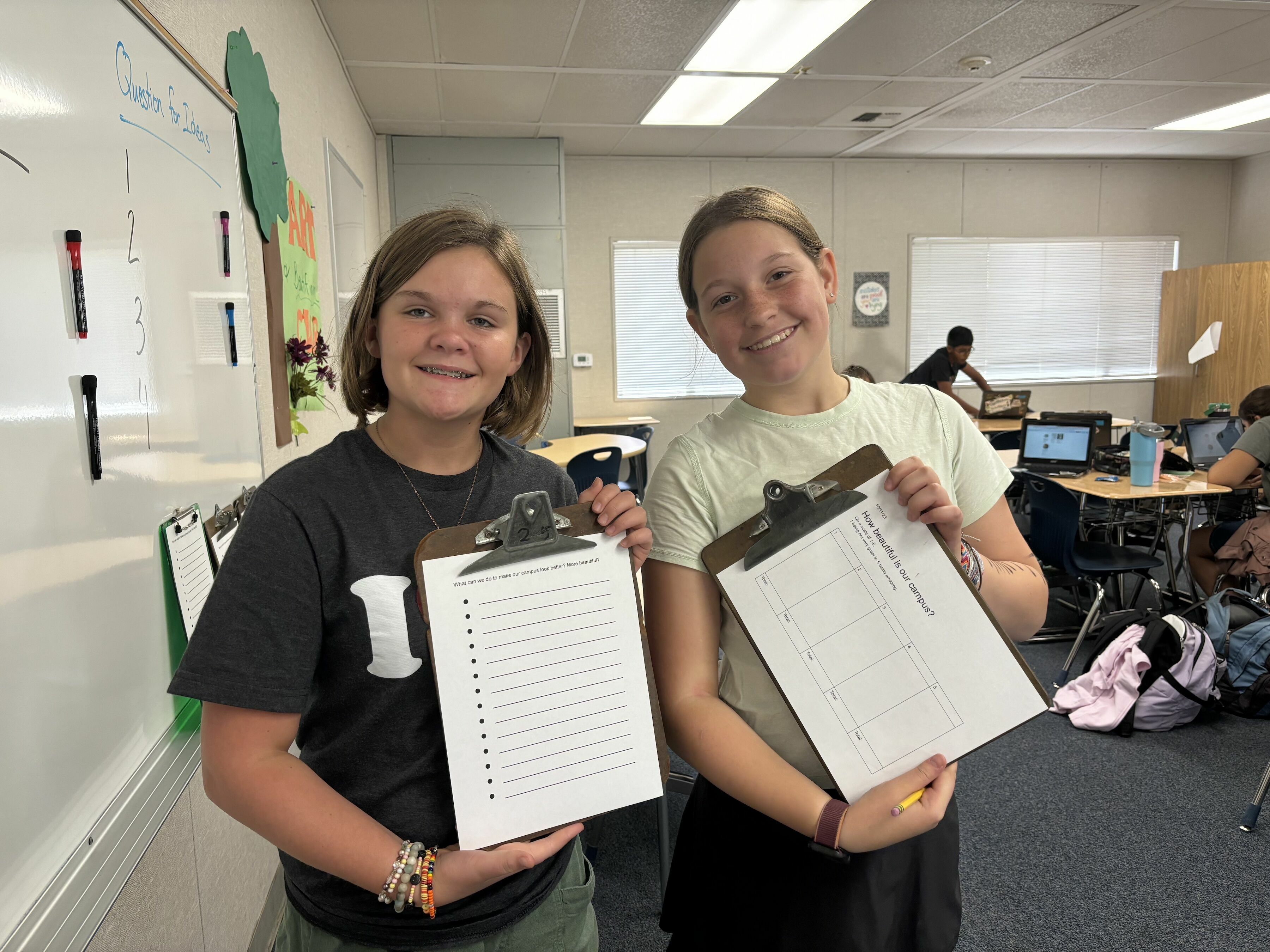 two pre-teen girls standing beside each other, smiling, and holding clipboards showing the forms they used to collect feedback from fellow students about campus beautification